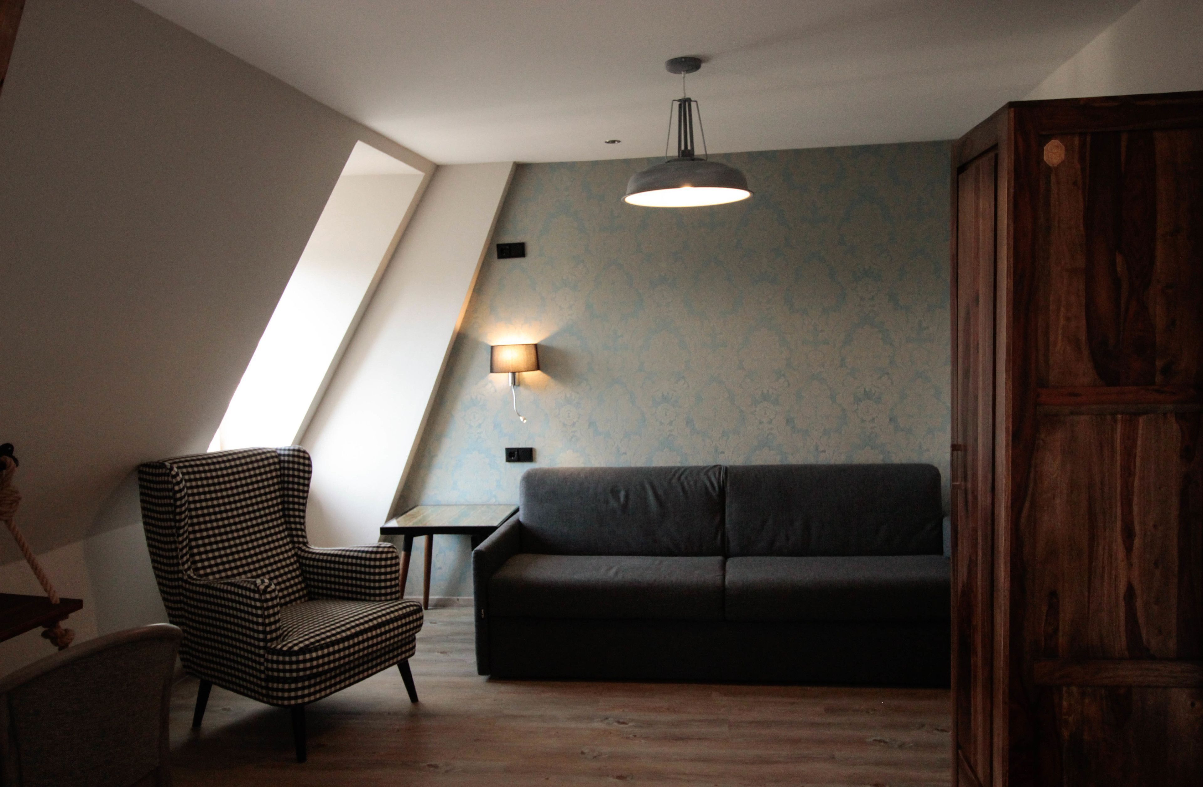 A loft-style room with a sloped ceiling, a black sofa, a checkered armchair, a small table, and a wall-mounted lamp.