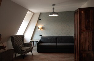 Quarto estilo loft com teto inclinado, sofá preto, poltrona xadrez, mesa pequena e luminária fixada na parede.
