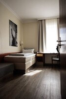Habitación de hotel con cama, escritorio, silla, radiador y ventana con cortinas.