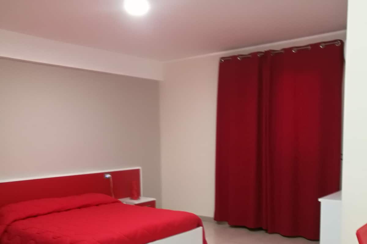 Ein Schlafzimmer mit einem weißen Bett, roten Vorhängen und einem weißen Nachttisch.