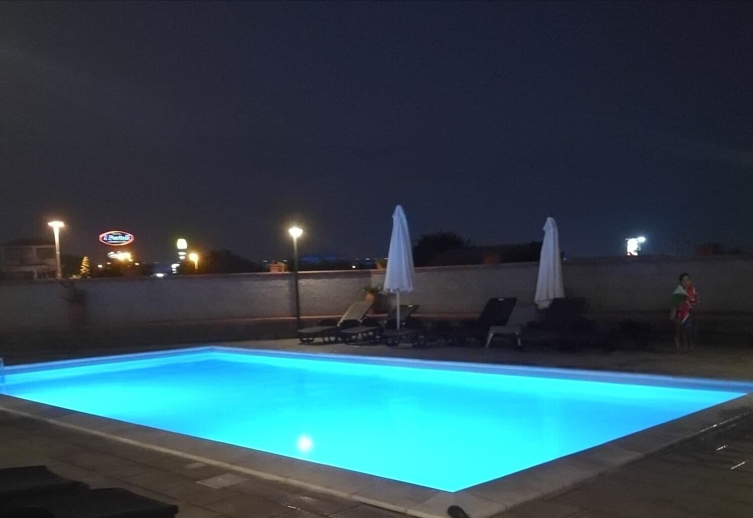 Ein Swimmingpool mit Liegestühlen und Sonnenschirmen, umgeben von einer niedrigen Mauer und Straßenlaternen.