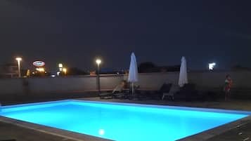 Ein Swimmingpool mit Liegestühlen und Sonnenschirmen, umgeben von einer niedrigen Mauer und Straßenlaternen.