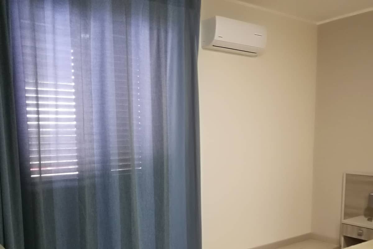 Ein Zimmer mit einem Fenster, das mit einem blauen Vorhang verdeckt ist, einer weißen Klimaanlage, die an der Wand montiert ist, und einem Bett mit weißer Bettdecke.
