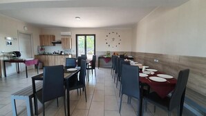 A spacious dining area with a long table set for multiple people, a kitchen area with wooden cabinets, and a large wall clock.
