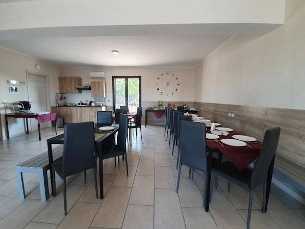 Een ruime eethoek met een lange tafel gedekt voor meerdere personen, een keuken met houten kasten, en een grote wandklok.