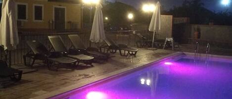 Piscina com espreguiçadeiras e guarda-sóis, cercada por um edifício com várias janelas e uma varanda.
