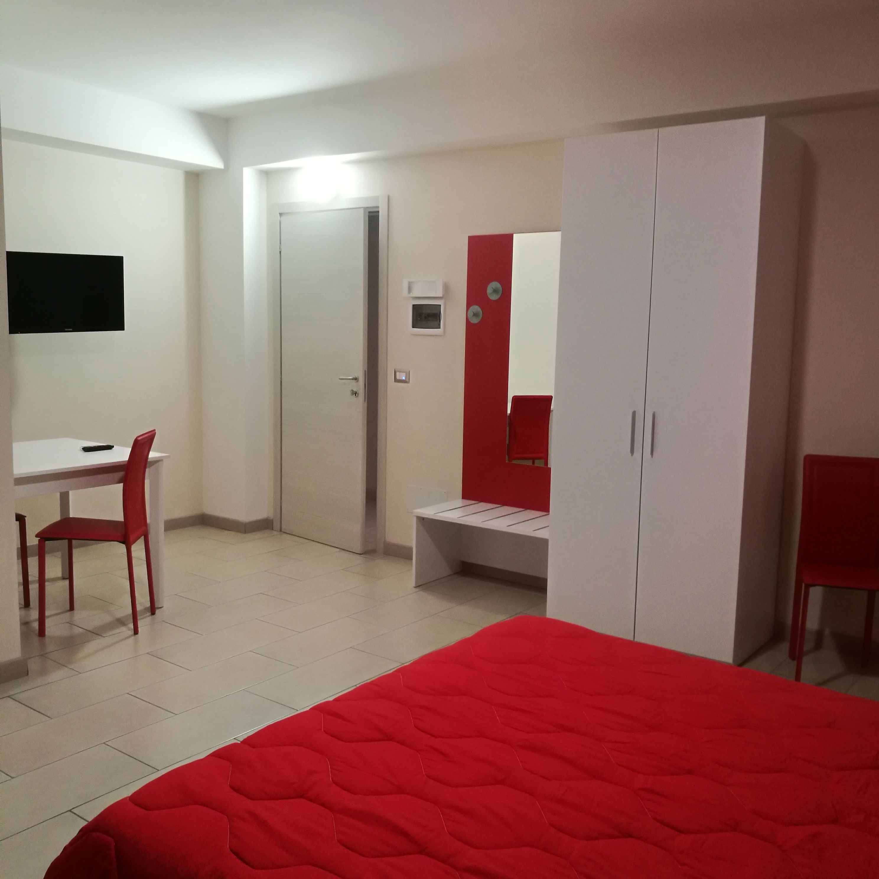 Una camera da letto con una coperta rossa, una scrivania con una sedia, un armadio e una televisione a parete.