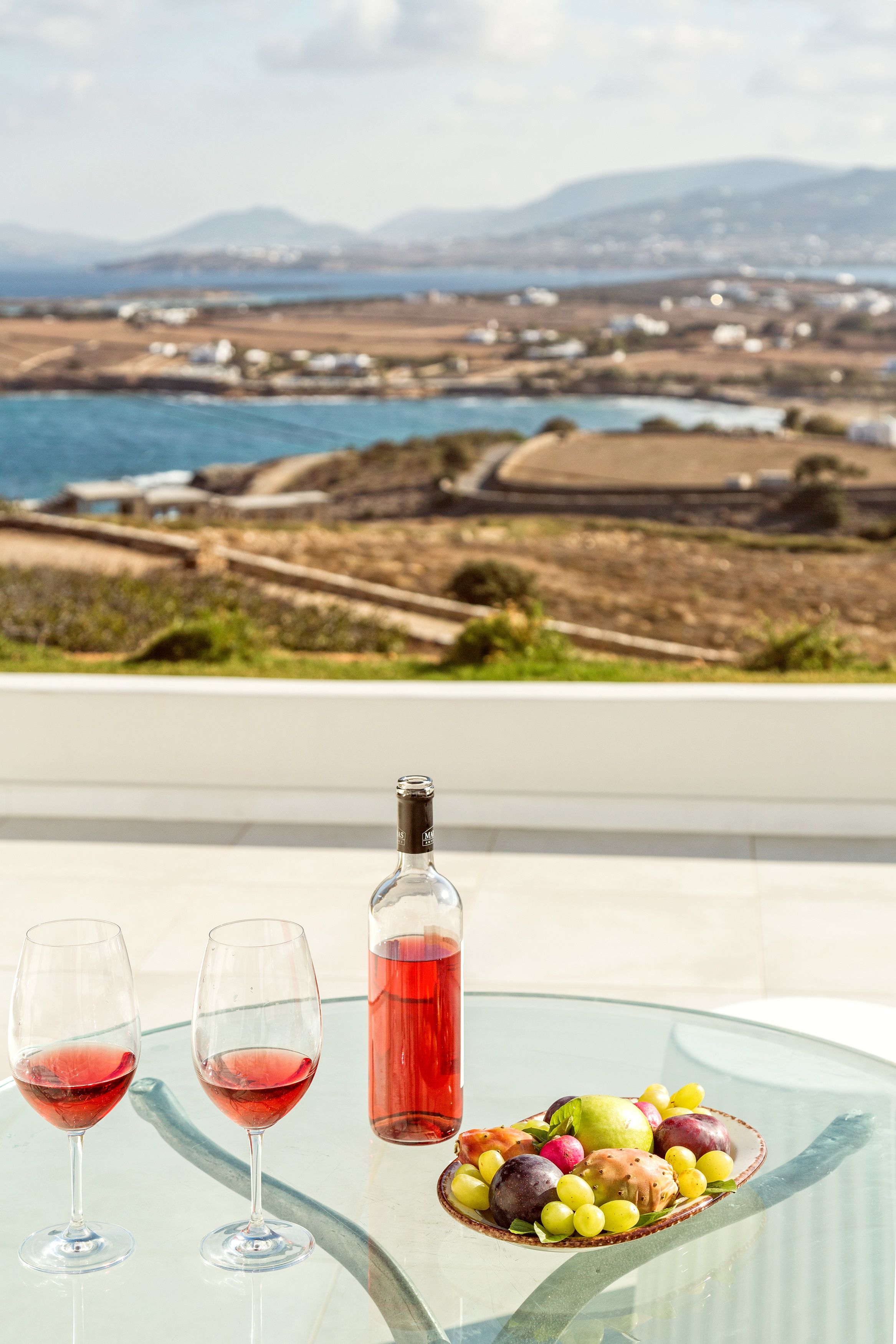 Una mesa con dos copas de vino, una botella y un frutero con uvas, melocotones y una pera.