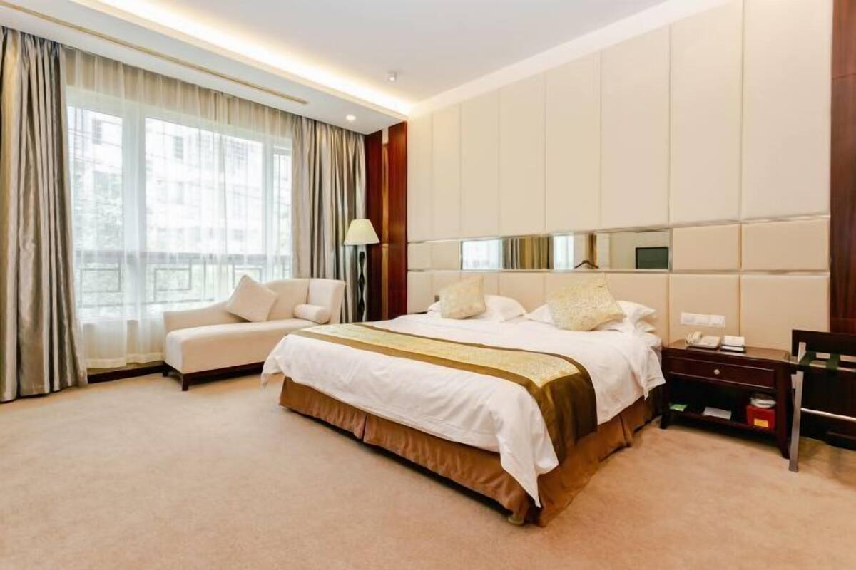 Une chambre d’hôtel avec un grand lit, un canapé, une table de chevet avec une lampe et une grande fenêtre avec des rideaux.