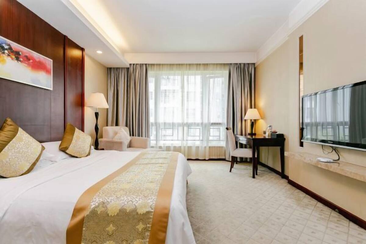 Une chambre d’hôtel avec un grand lit, un bureau, une chaise, une télévision et une fenêtre avec des rideaux.