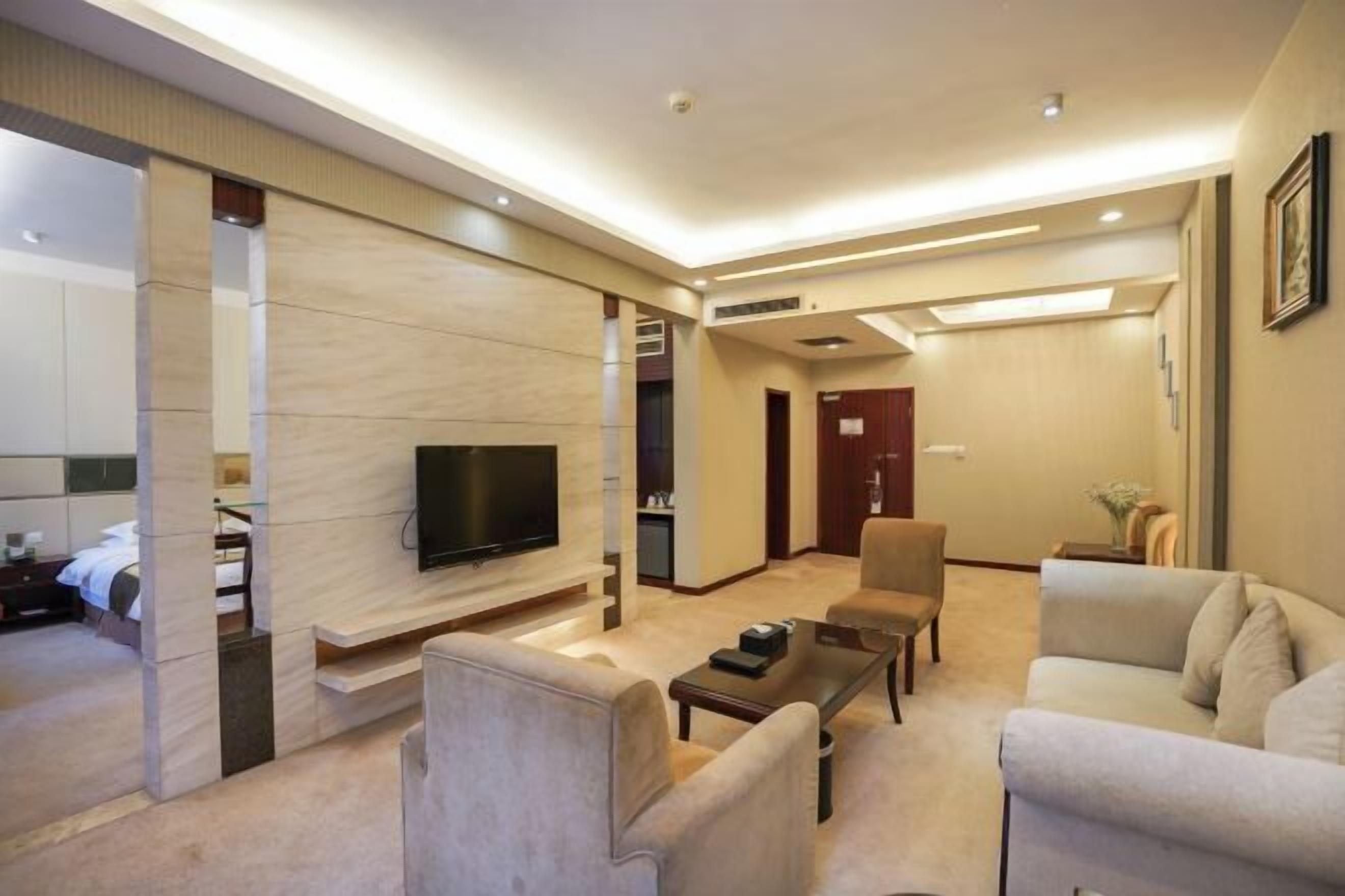 A modern hotel room with a flat-screen TV mounted on a wall, a sofa, a coffee table, and a separate bedroom area with a bed and a nightstand.