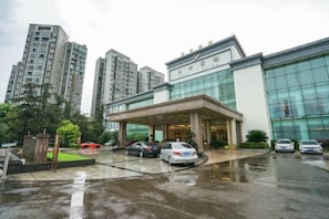 A modern hotel entrance with a covered driveway, tall surrounding buildings, and parked cars.