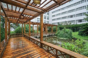 Eine Holzterrasse mit Blick auf einen Teich und viel Grün, mit Ausblick auf ein mehrstöckiges Gebäude.