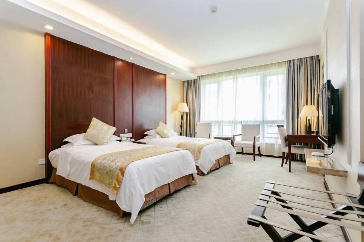 Une chambre d’hôtel avec deux lits, un bureau, une chaise, une télévision et une grande fenêtre avec des rideaux.