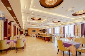 Un amplio comedor de hotel con numerosas mesas y sillas, un bar central y grandes ventanales.