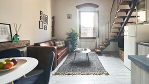 A modern living room with a brown leather sofa, a dining table with fruits, a staircase, and a kitchen area. - A Few Steps to The Duomo Adorable 1BD Apt (Florence)