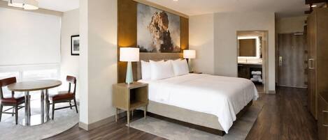 A hotel room with a bed, bedside table, lamp, chair, and a painting on the wall.