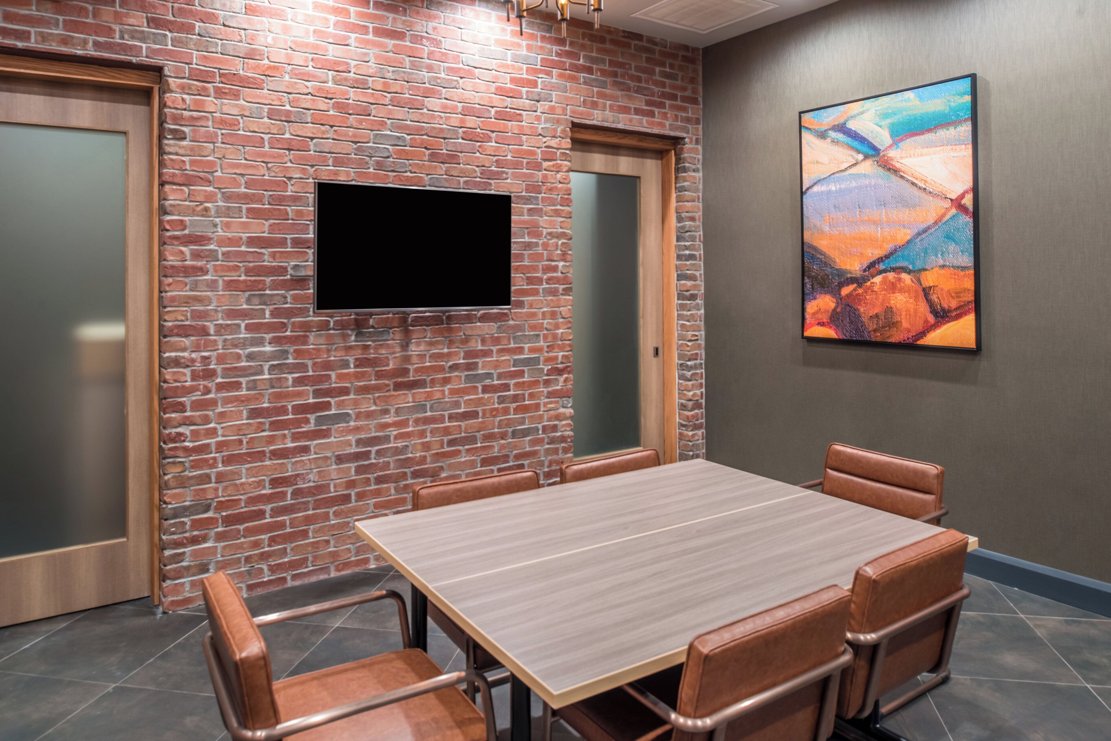 Een vergaderruimte met een bakstenen muur, een flatscreen televisie, een schilderij en een houten tafel met lederen stoelen.