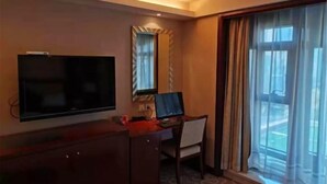 A hotel room with a TV, a desk with a computer, a mirror, and a large window with curtains. - Jinling Netda Hotel (Nantong)