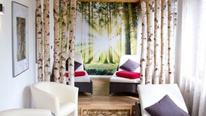 A room with a wooden ceiling, birch tree wall decor, two white lounge chairs with red and black cushions, and a large window with curtains. - Kainsbacher Mühle (Happurg)