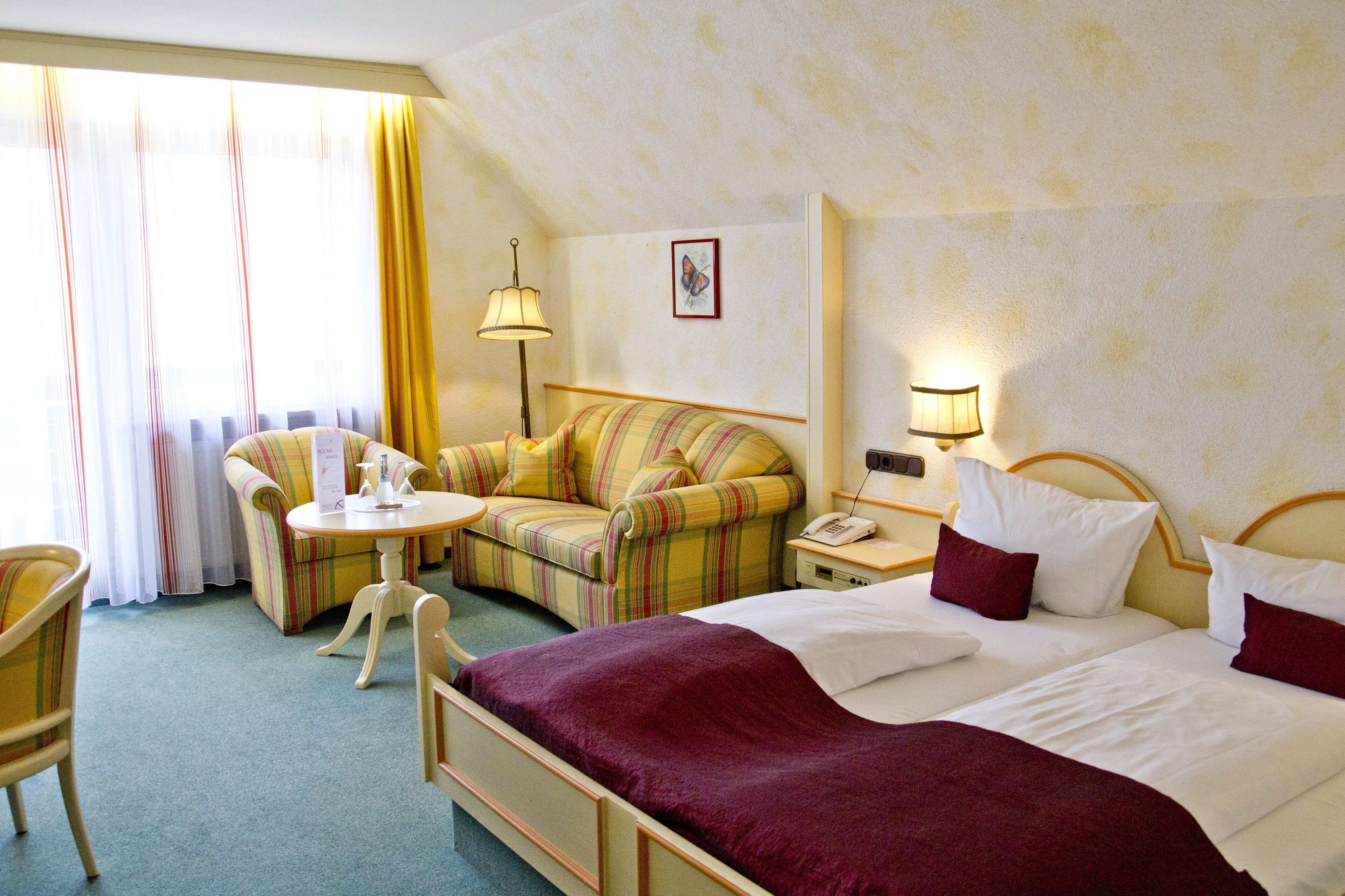 A hotel room with a bed, a sofa, a small table, and a chair. There is a window with curtains, a lamp, and a wall-mounted phone.