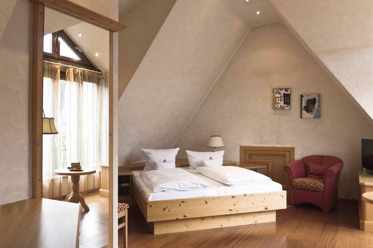 A bedroom with a sloped ceiling, a wooden bed, a chair, a table, and a lamp.