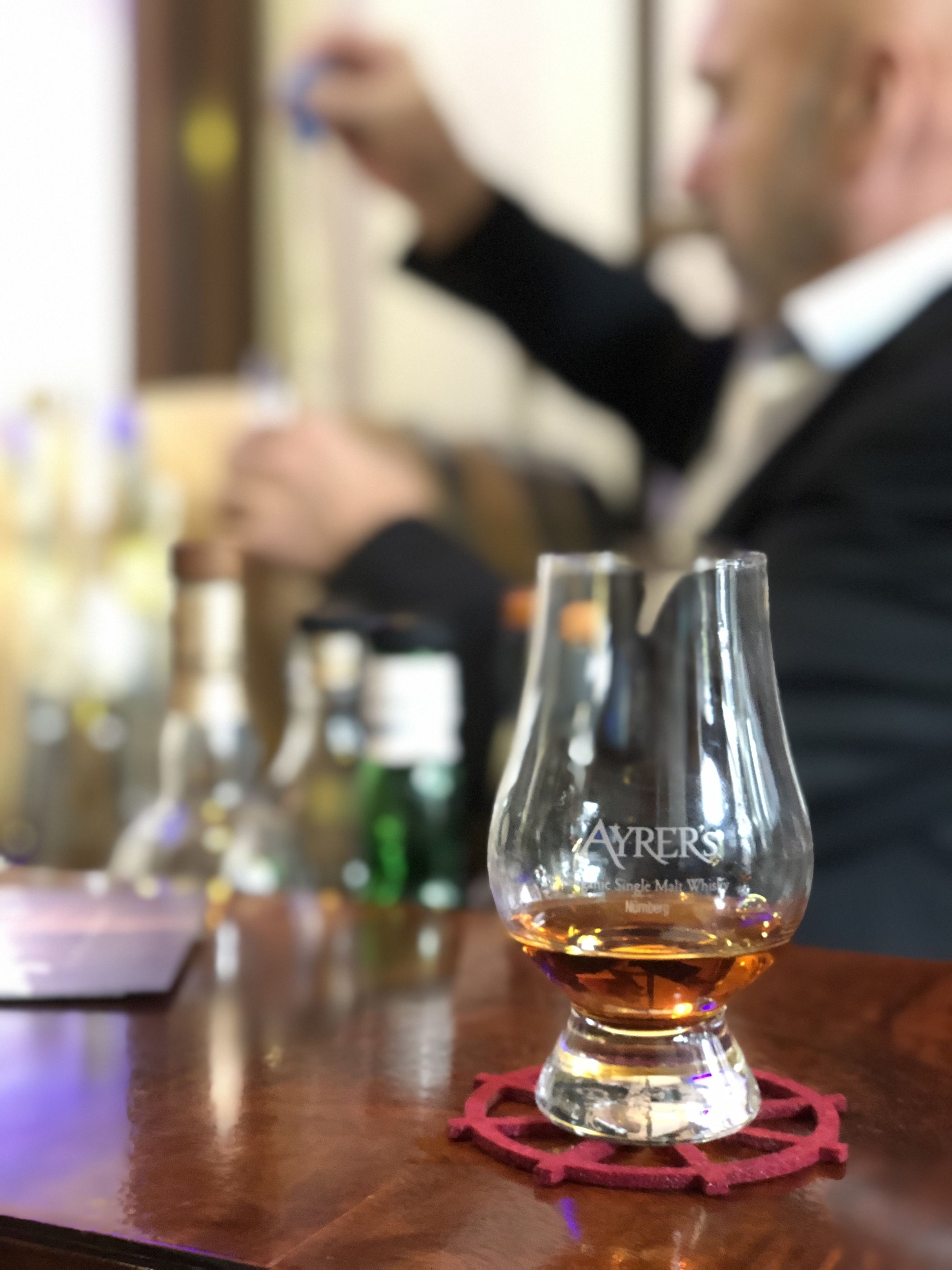 Un vaso de whisky sobre una barra de madera con un posavasos rojo, y un fondo borroso con una persona y botellas.