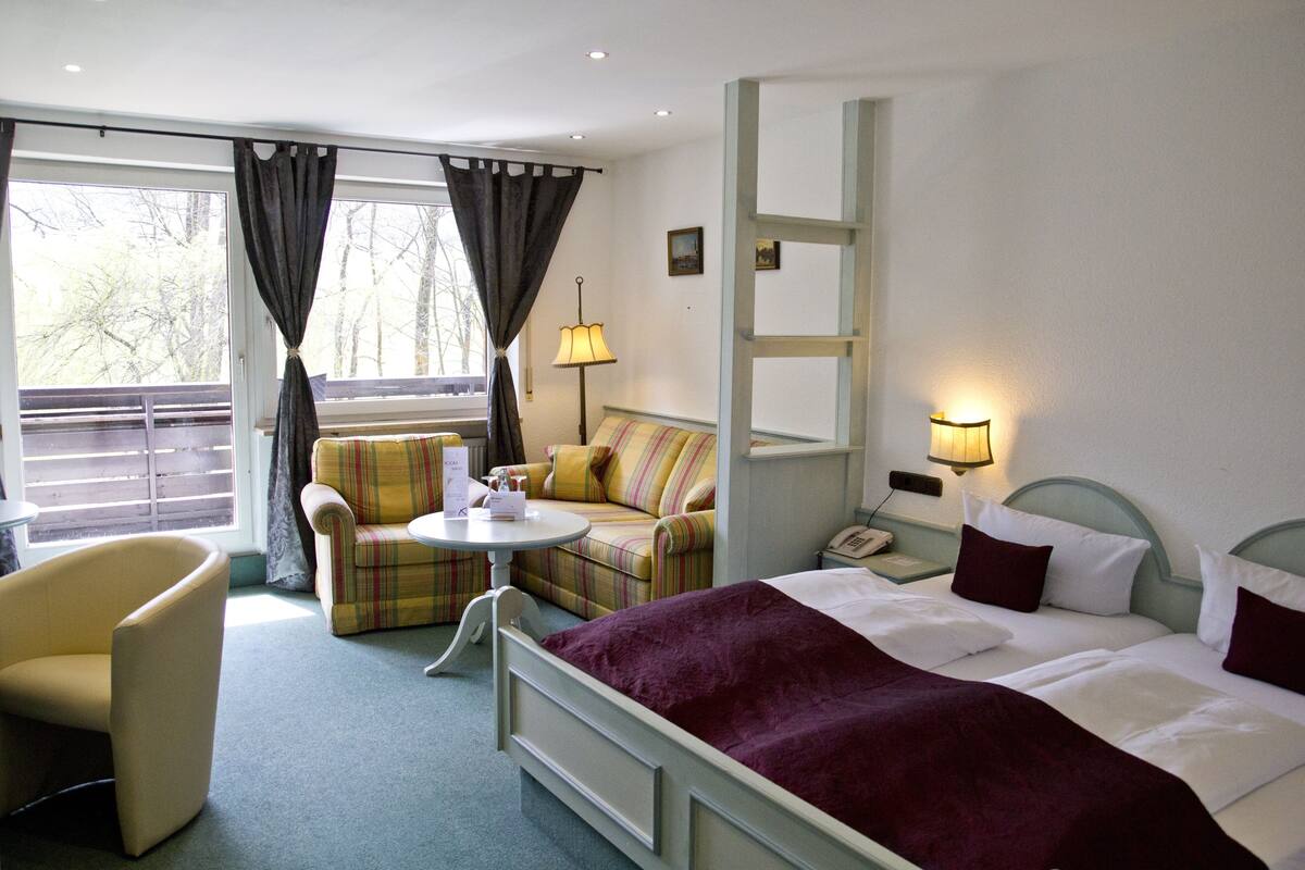 A hotel room with a bed, a sofa, a chair, a small table, and a nightstand with a lamp.