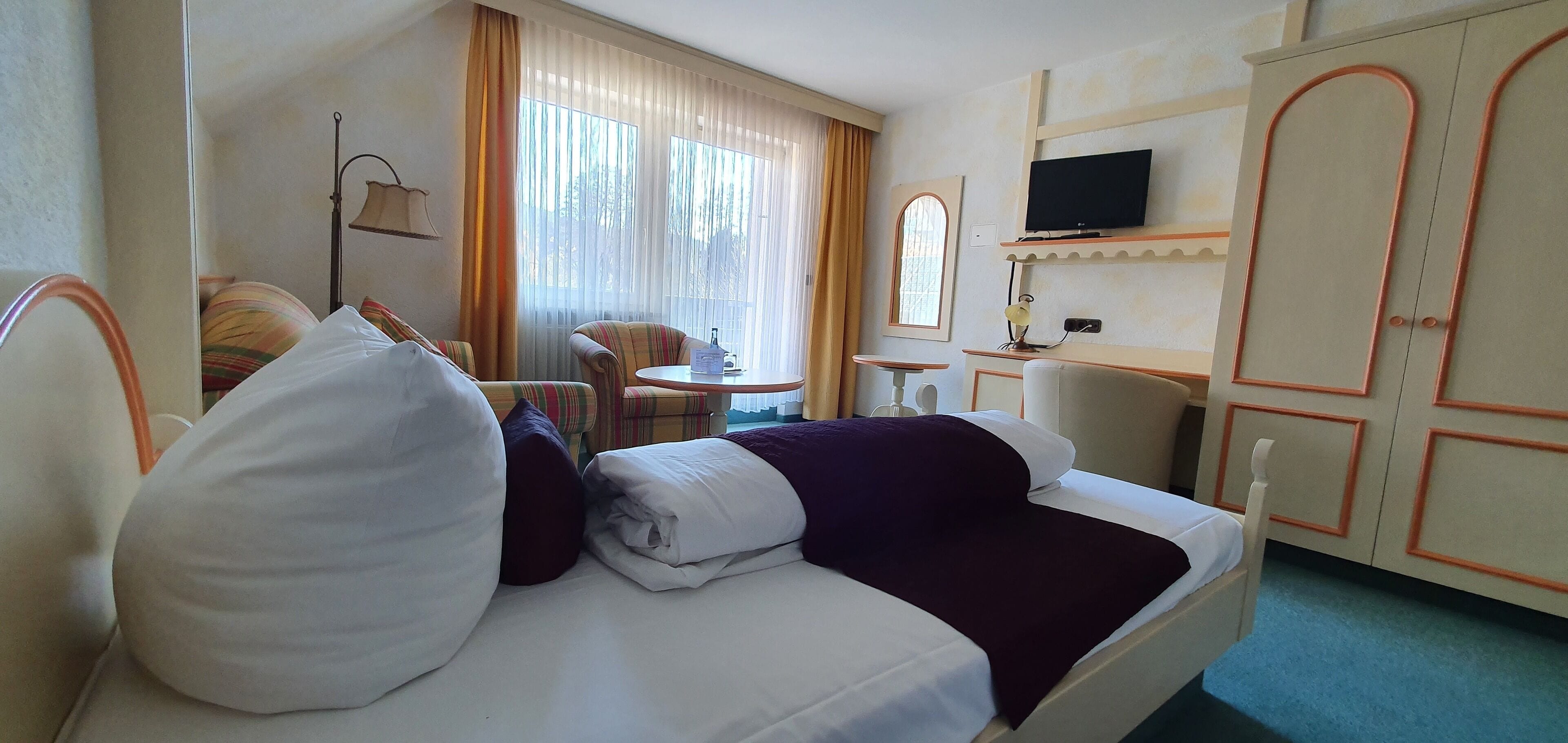 A hotel room with a bed, a small table, a chair, a TV, and a wardrobe.