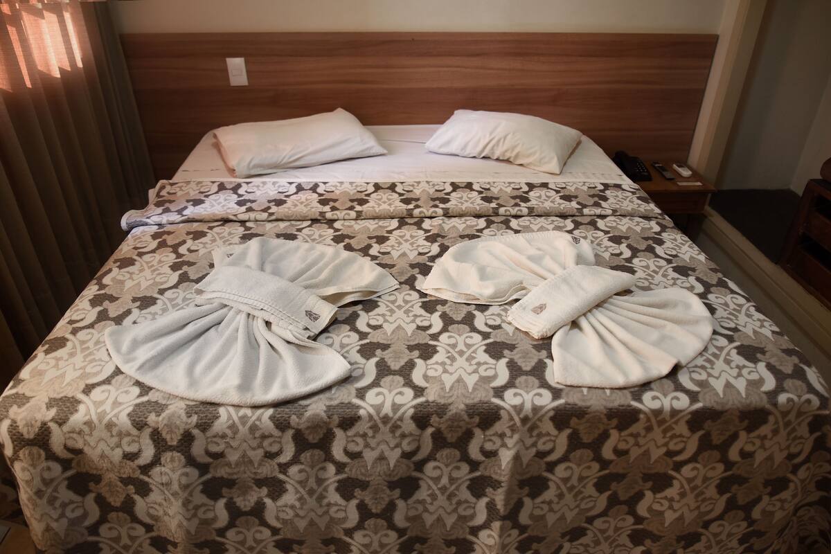 A hotel room with two beds, each with a patterned bedspread and pillows. There is a wooden headboard, a nightstand with a phone and remote control, and a window with curtains.