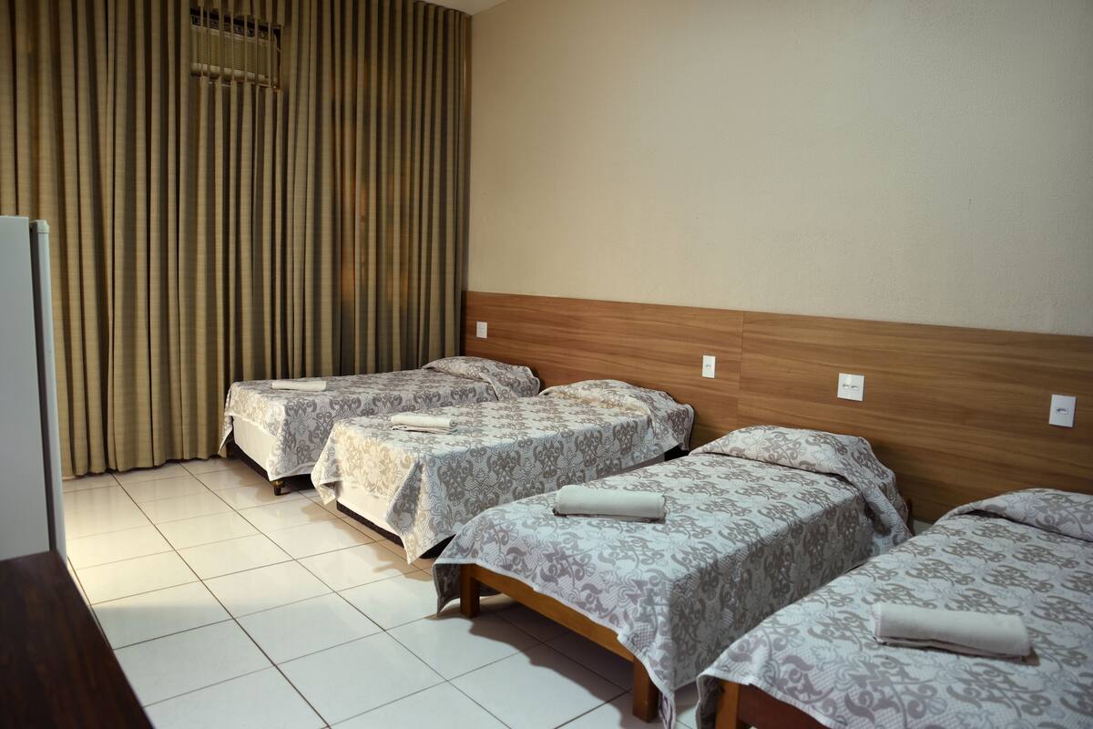 A hotel room with three single beds, a wooden headboard, a large window with vertical blinds, and a tiled floor.