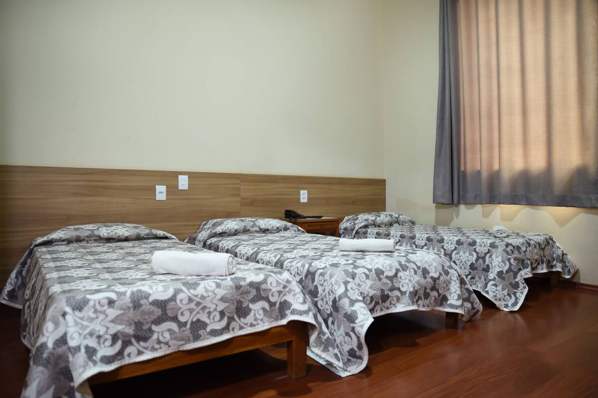 Two single beds with patterned bedding, wooden headboards, a bedside table with a lamp, and a window with curtains.