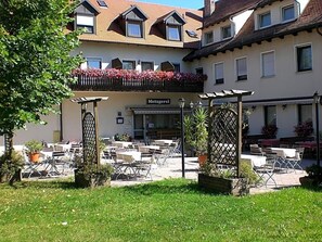 An outdoor seating area with tables and chairs, a building with a sign reading 'Metzgerei,' and a garden with potted plants.