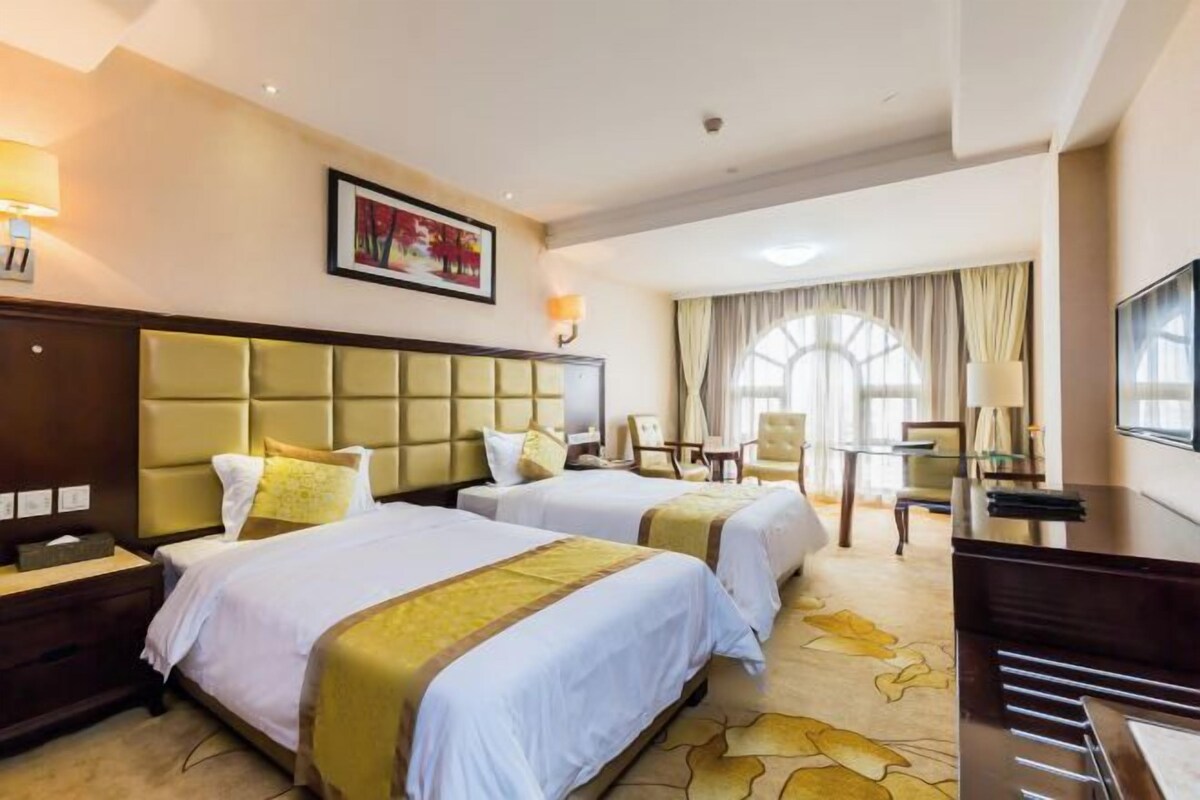 A hotel room with two beds, a desk, a chair, a TV, and a large window.