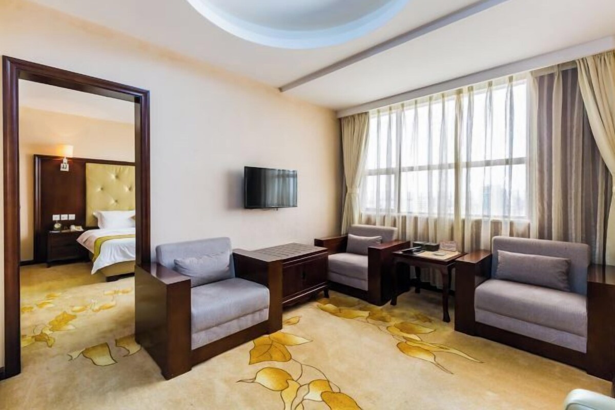 A hotel room with a bed, two armchairs, a small table, a TV, and a large window with curtains.