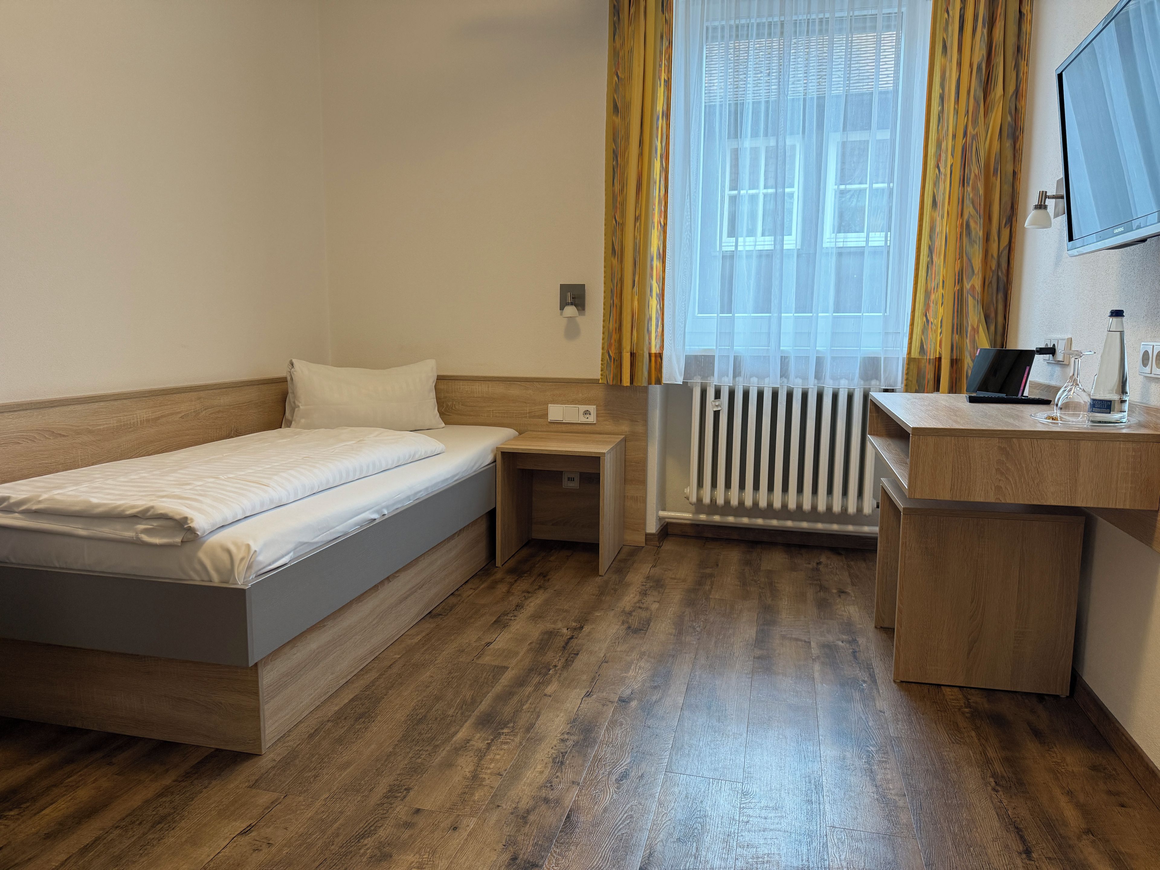 A hotel room with a single bed, a desk, a chair, a radiator, and a window with curtains.