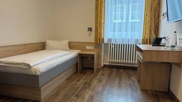 A hotel room with a single bed, a desk, a chair, a radiator, and a window with curtains.