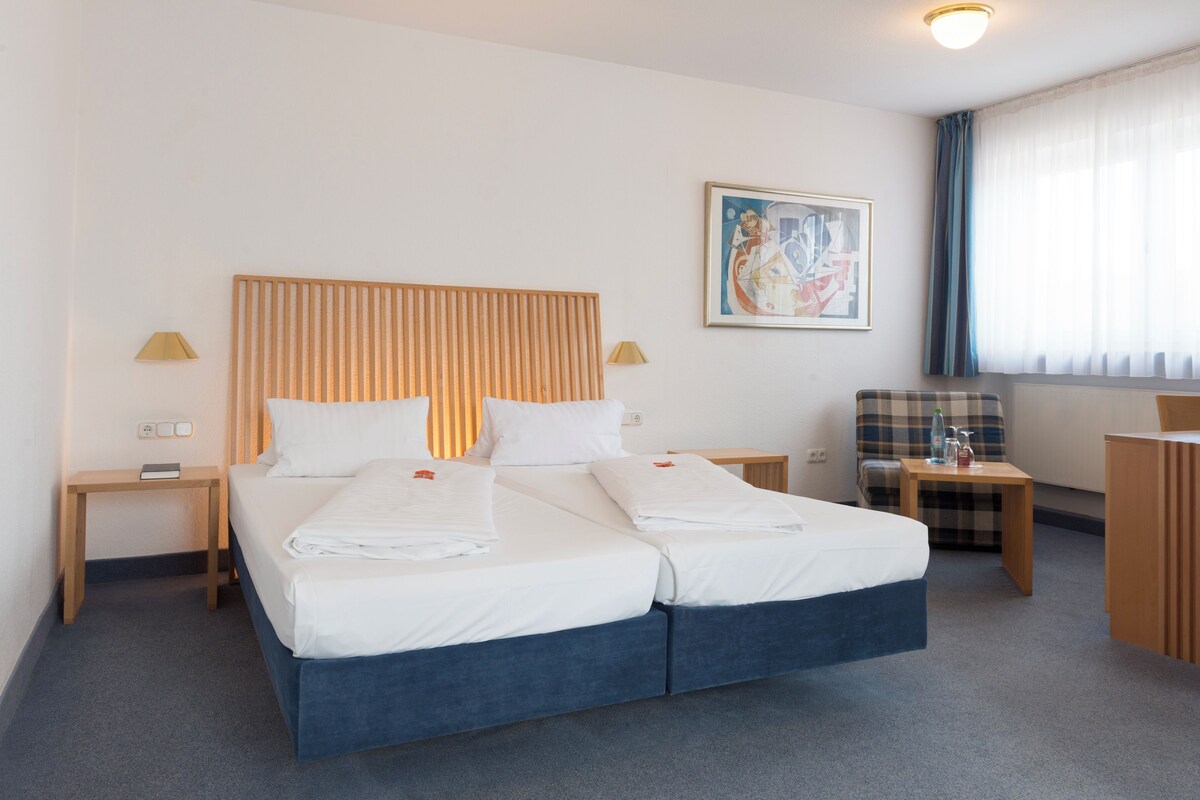 A hotel room with a double bed, a bedside table with a lamp, a chair, a small table with a bottle and glass, and a painting on the wall.