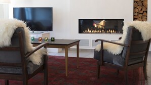 Ein Wohnzimmer mit Flachbildfernseher, Kamin, zwei Sessel mit Felldecken und einem roten Teppich mit Muster.