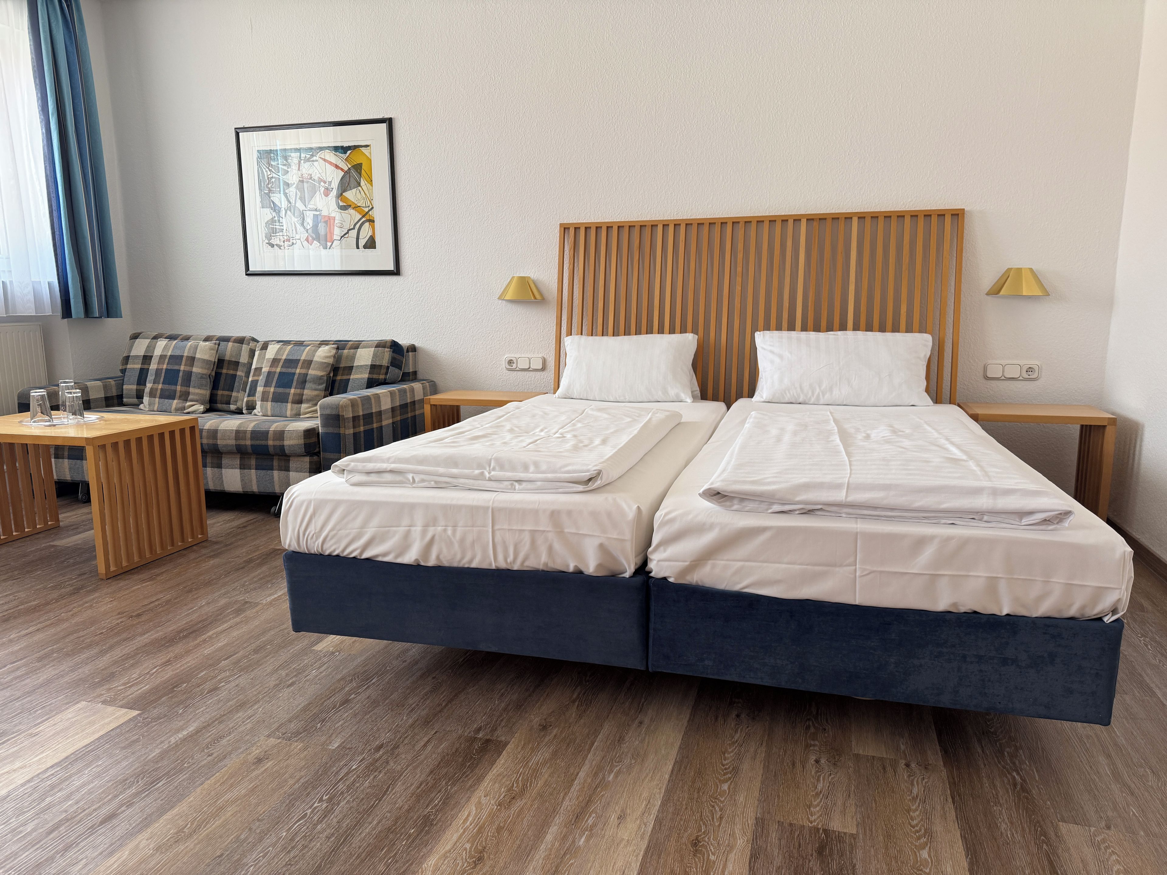 Een tweepersoonsbed met houten hoofdbord, een klein nachtkastje, een lamp en een bank met ruitkussens.