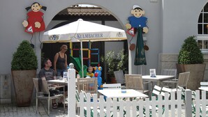 Ein Außenbereich mit Café, Tischen, Stühlen und Sonnenschirm, sowie Spielgeräte für Kinder.
