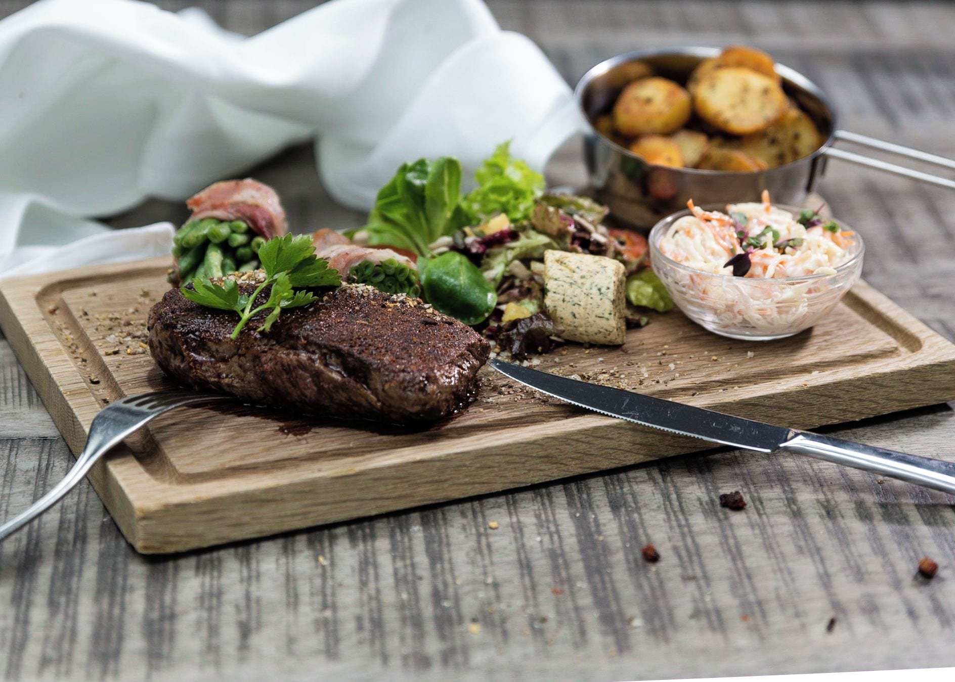 A wooden cutting board with a steak, a salad, and a bowl of coleslaw.