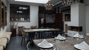 Un'elegante sala da pranzo moderna con un grande tavolo in legno apparecchiato per sei persone, circondato da sedie nere. Sopra, un lampadario, e sulla destra si intravede un camino.