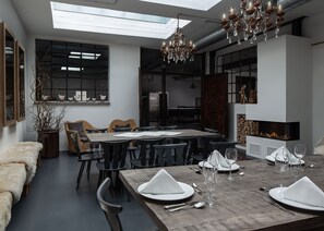 A modern dining area with a large wooden table set for six, surrounded by black chairs. A chandelier hangs above, and a fireplace is visible on the right.
