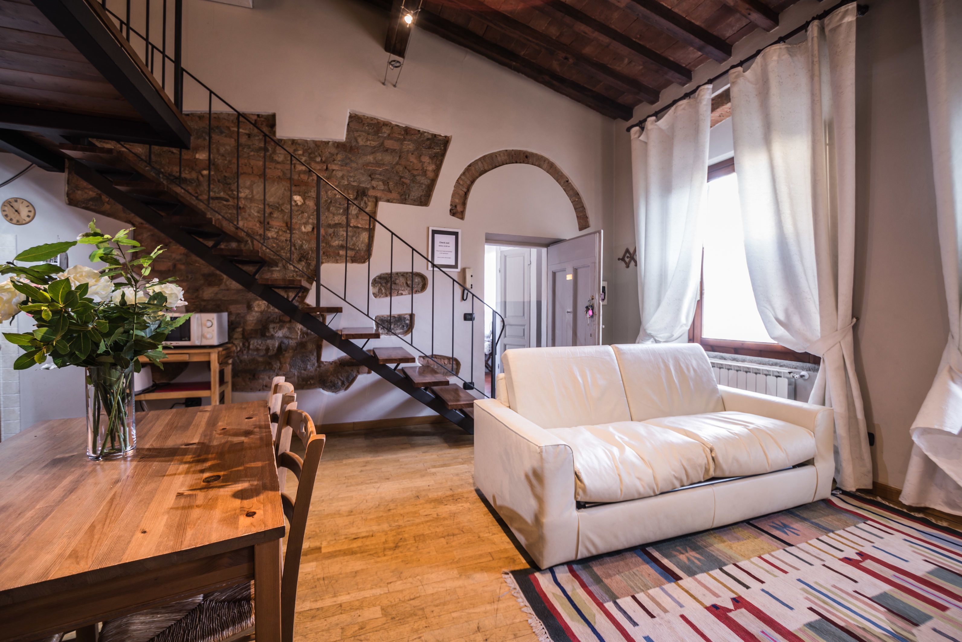 A cozy living room with a wooden dining table, a white sofa, and a staircase with stone walls.