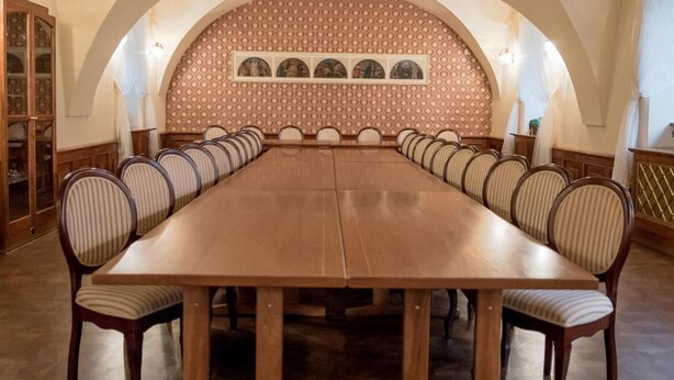 Una larga mesa de comedor con sillas tapizadas, techo abovedado, pared con diseño y una obra de arte enmarcada encima.