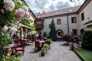 An outdoor patio with tables and chairs, surrounded by greenery and a building with a tiled roof.