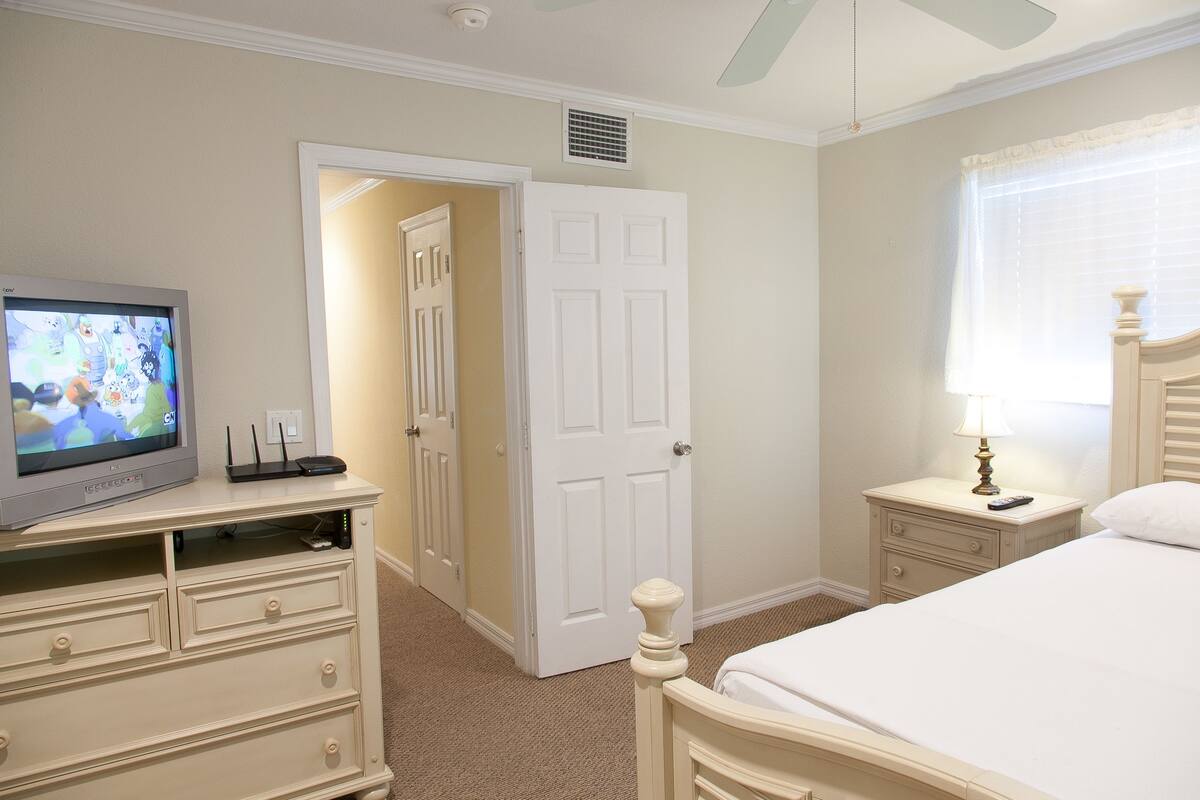 Une chambre à coucher comprenant un lit, une table de chevet, une commode, un téléviseur et une porte donnant sur une autre pièce.