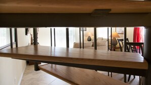 Una panca di legno con struttura in metallo in una stanza luminosa, con un divano, una lampada e un tavolo da pranzo.