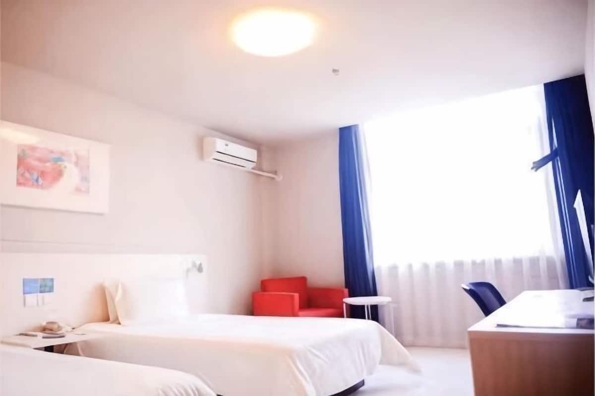 A hotel room with two beds, a desk, a chair, a red armchair, and a window with curtains.